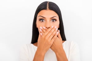 Shocking news. Surprised young woman covering mouth with hands and staring at camera while standing against white background