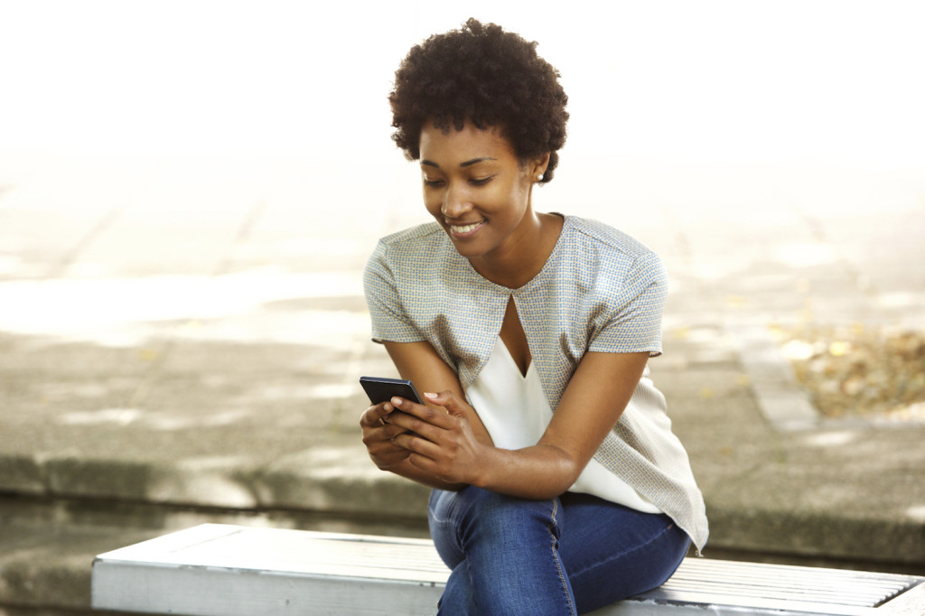 Portrait of happy young african woman sitting outside on bench reading a text message on her mobile phone