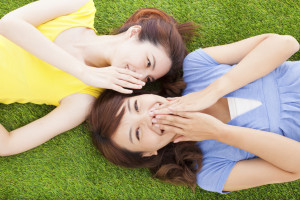 Two people lying in the grass. One whispers into the ear of the other, who has their hands over their mouth and seem to be laughing.