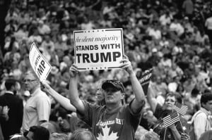A black and white photo of a rally, with an individual in a baseball cap holding the sign, "The silent majority stands with Trump."