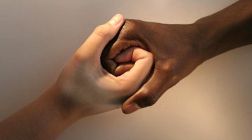 Two hands – one with a light skin tone, one with a dark skin tone – are holding each other.