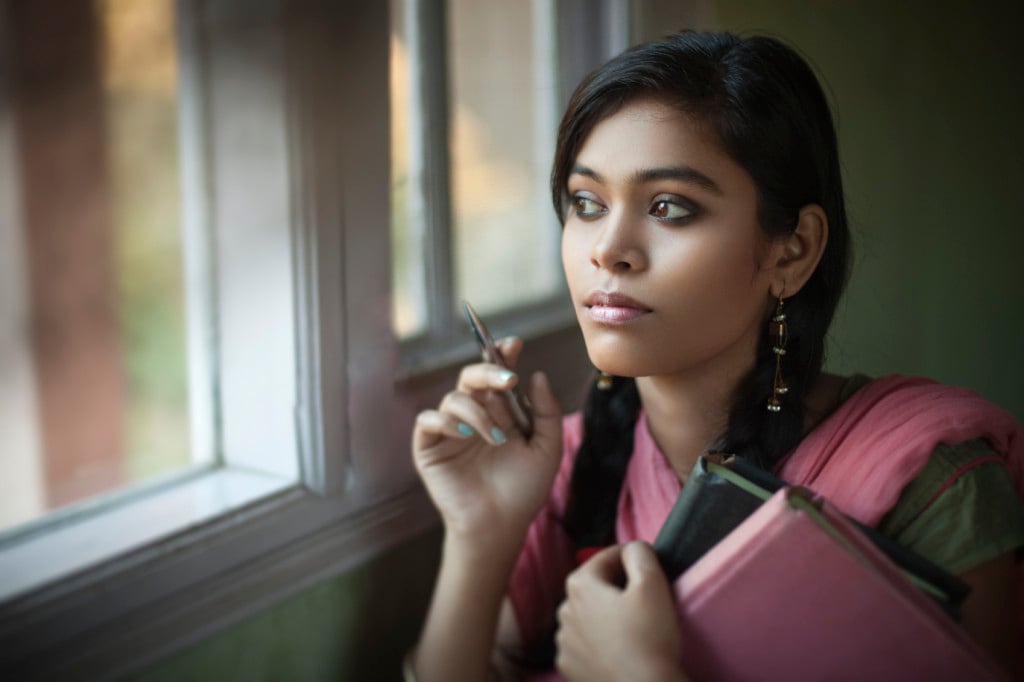 A young person is standing by a window in a school, looking out serenely. They are holding pink and black notebooks and a pen. They are wearing a pink and green sari.