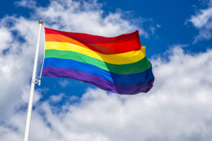 A rainbow flag flying in the wind with a cloudy blue sky in the background.