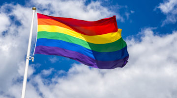 A rainbow flag flying in the wind with a cloudy blue sky in the background.