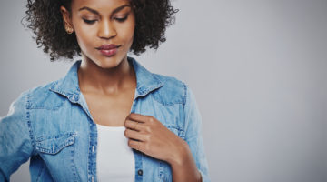 A person appearing thoughtful as they stand, looking down, tugging at their denim coat with one hand.
