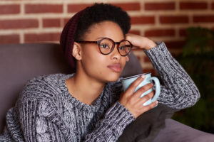 A young person sits on a couch, supporting their head with one hand and holding a mug with the other. They appear thoughtful.