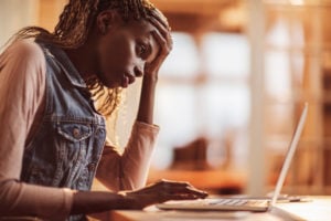 A person sits in front of a laptop, appearing worried, with one hand touching their forehead.