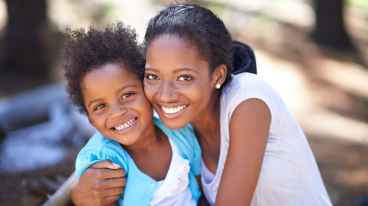 A parent hugging a small child in nature. Both smile happily as they look into the camera.