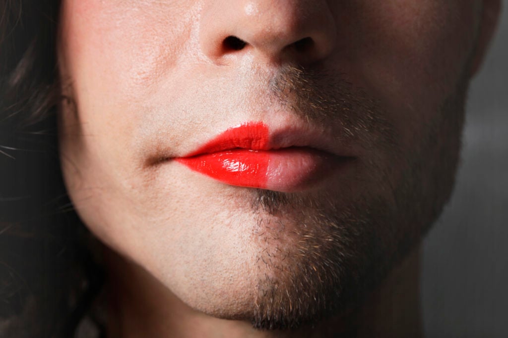 Closeup of the lower half of a face. One half is clean shaven and has long hair and bright red lipstick; the other half is unshaven with short hair and no lipstick.