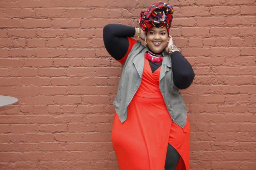 Black Muslim woman posing with her hands behind her head against a brick wall, smiling at the camera.