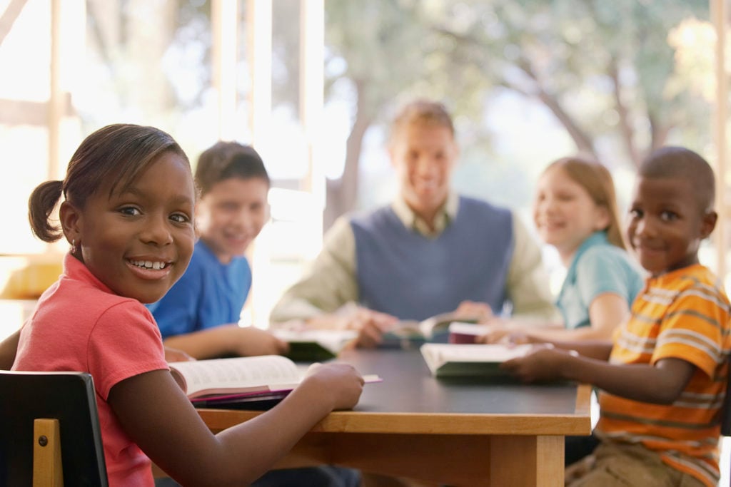 A group of students sit at a table with their teacher, reading and smiling. The photo has a shallow depth of field.
