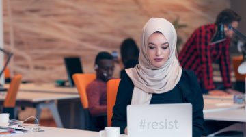 A person wearing a hijab sits at their desk with coworkers in the background. Their laptop reads "#resist." Image source: Thinkstock; text added by Everyday Feminism.