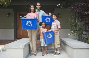 A smiling family of four stand in front of their house, holding recycling bins.