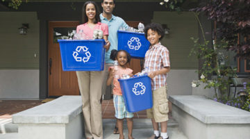 A smiling family of four stand in front of their house, holding recycling bins.