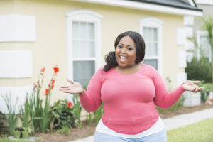 A fat person stands in front of their house, smiling and raising their hands to shoulder level.
