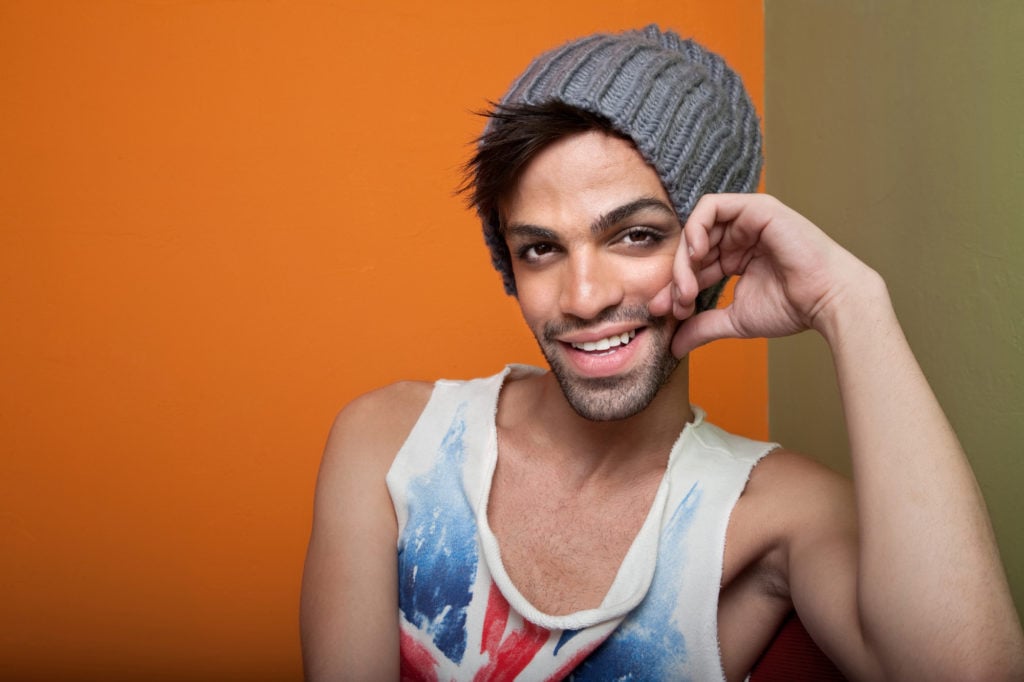 A person wearing a gray beanie and a tank top smiles, their hand resting against their cheek.